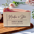 Handcrafted cold process soap in a box labeled 'Meadow & Stone Dewy Rose' with flowers in the background.