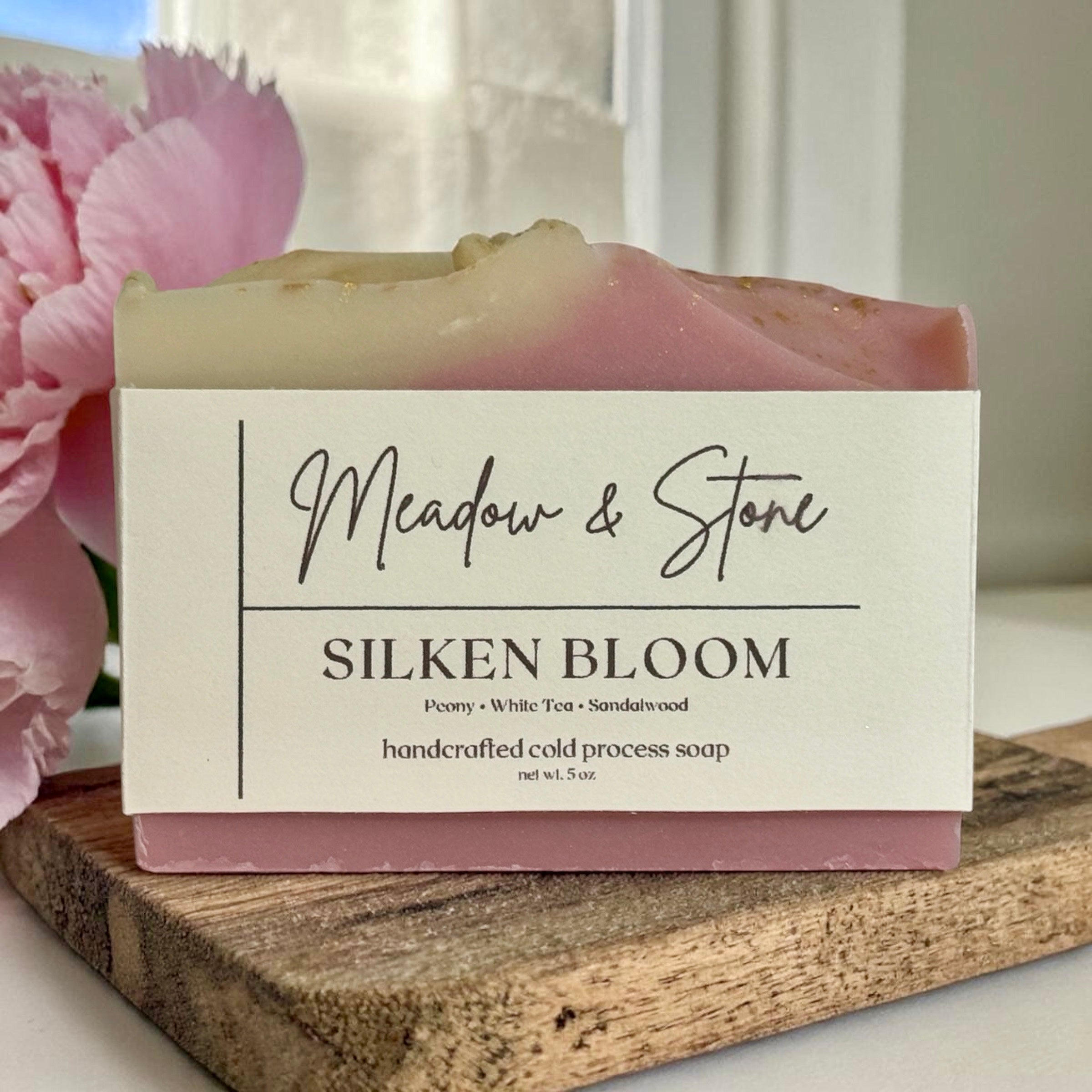 Bar of soap with 'Meadow & Stone Silken Bloom' label on a wooden block with pink flowers in the background.